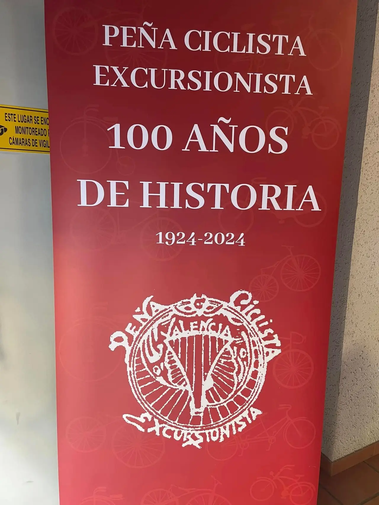 Cartel vertical del club, en rojo, con el texto «Peña Ciclista Excursionista, 100 años de historia, 1924-2024» y el emblema debajo