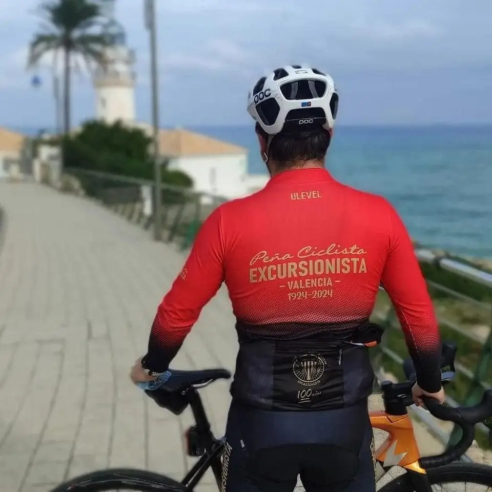 Un socio de espaldas, apoyado en su bicicleta frente al mar, con el maillot del centenario en el que se lee «Peña Ciclista Excursionista · Valencia · 1924-2024»