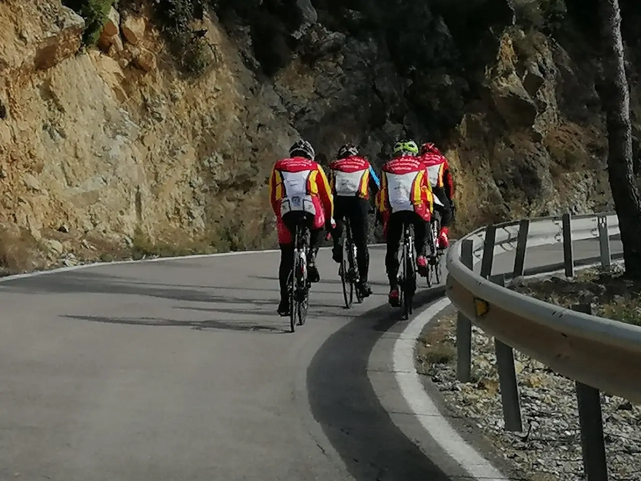 Cuatro socios con el maillot rojo y amarillo del club suben en fila una carretera de montaña, vistos desde atrás, entre una pared de roca y el quitamiedos