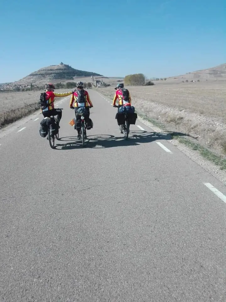 Tres cicloturistas del club con alforjas ruedan de espaldas por una carretera recta de meseta, con un cerro coronado por un castillo al fondo