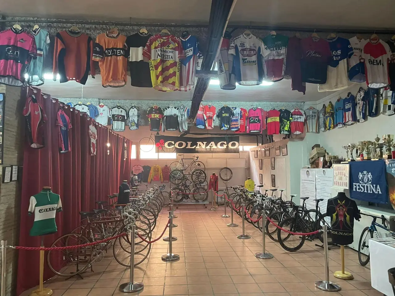 Sala del museo con una veintena de bicicletas antiguas acordonadas, decenas de maillots colgados del techo y una vitrina de trofeos